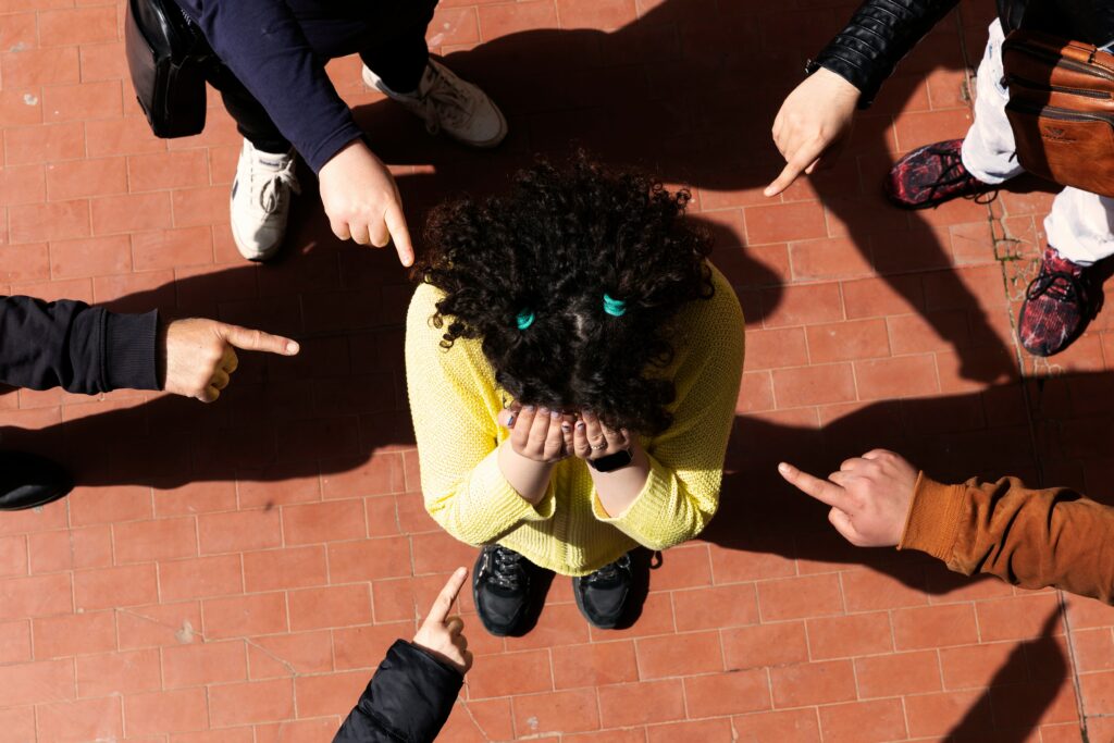 A person with their head in their hands, being pointed at by multiple fingers, visually representing the feeling of shame and isolation during bullying.
