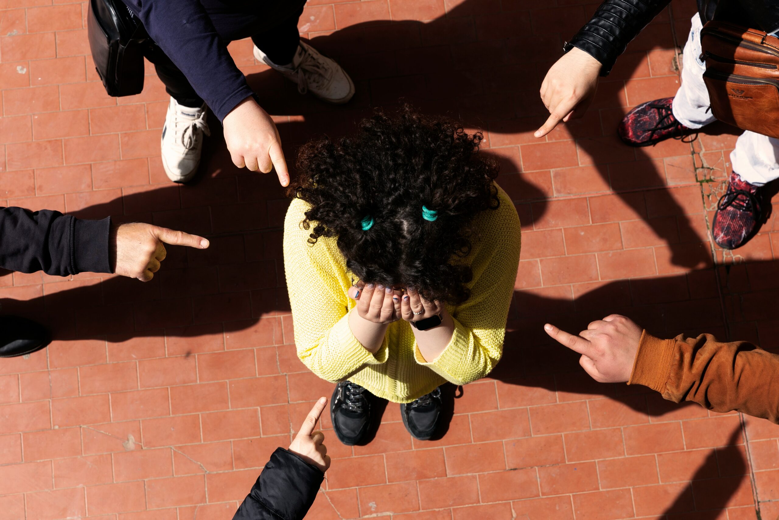 A person with their head in their hands, being pointed at by multiple fingers, visually representing the feeling of shame and isolation during bullying.